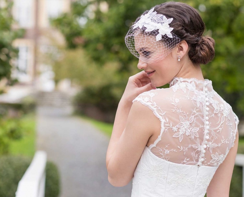 Braut in weißem Spitzenkleid mit Knopfleiste am Rücken und Hochsteckfrisur mit Haarschmuck, steht im Freien