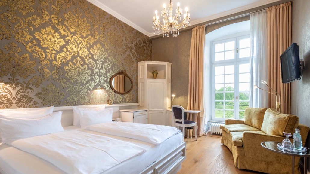 Doppelzimmer golden Doppelzimmer mit zwei Einzelbetten, goldgemusterten Tapeten, Kronleuchter, Schreibtisch, gelbem Sofa und großem Fenster mit Blick ins Grüne