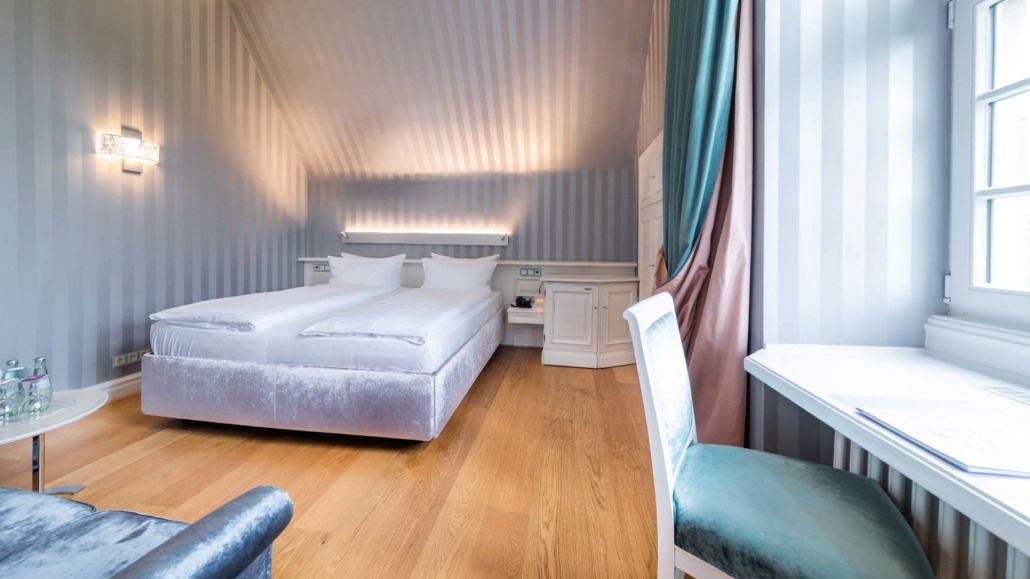 Doppelzimmer mit Blick auf das Bett Helles Doppelzimmer mit großem Bett, weißem Schreibtisch und Stuhl, Holzboden und Fenster mit Vorhängen