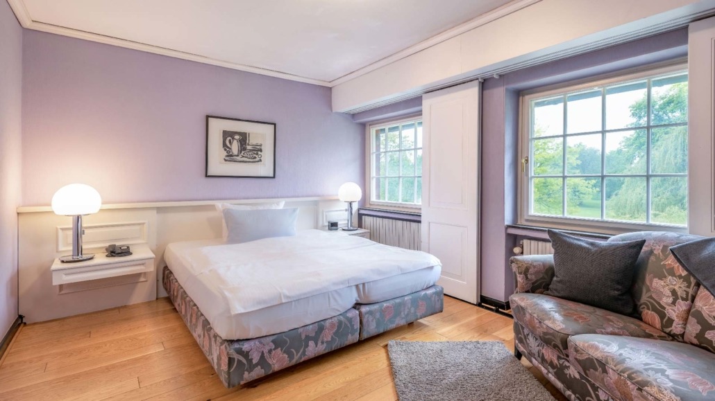 Einzelzimmer Einzelzimmer mit großem Bett, lila Wand, zwei Nachttischlampen, Sofa mit floralem Muster und zwei Fenstern mit Blick ins Grüne