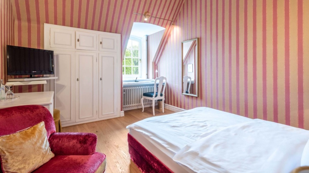 Einzelzimmer Dachgeschoss Einzelzimmer mit weißem Bett, rotem Sessel, weißem Schrank und Stuhl vor Fenster mit Blick nach draußen