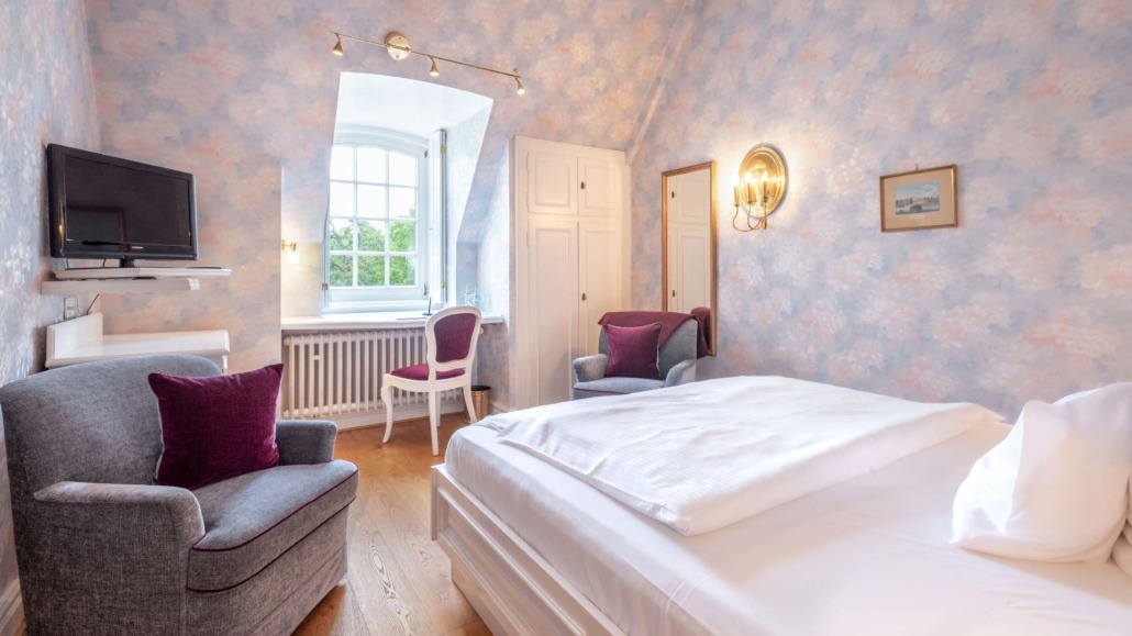 Kleineres Einzelzimmer Einzelzimmer mit weißem Bett, grauem Sessel mit rotem Kissen, Schreibtisch vor Fenster und hell gemusterter Wand