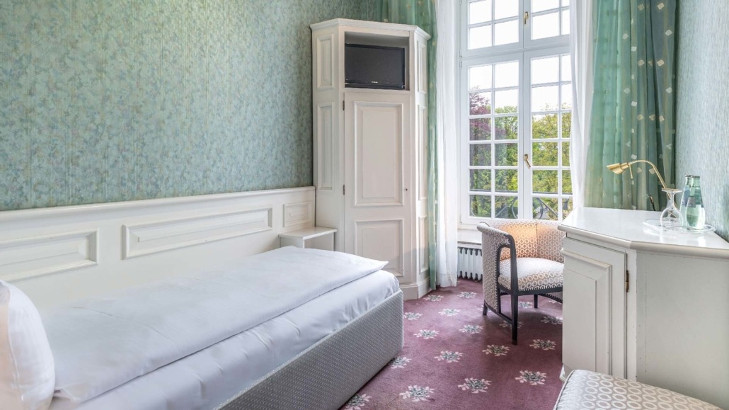 Einzelzimmer mit Einzelbett Einzelzimmer mit einem Bett, einem weißen Schrank mit Fernseher, einem Sessel am Fenster und einem Schreibtisch mit Wasserflasche