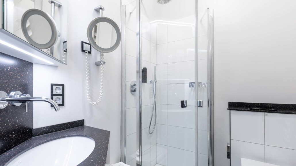 Einzelzimmer - Baderaum Modernes Badezimmer mit Waschbecken, beleuchtetem Spiegel, Duschkabine mit Glaswänden und weißen Fliesen