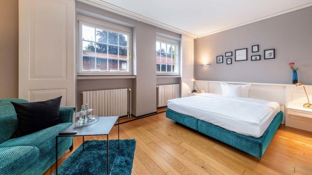 Einblick ins Einzelzimmer Einzelzimmer mit Doppelbett, blauem Sofa, zwei Fenstern und Holzboden