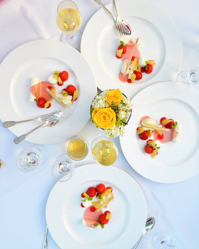 Vier Teller mit kunstvoll angerichteten Vorspeisen aus rohem Fisch, Erdbeeren und kleinen Gemüsestücken auf weißem Tischtuch, umgeben von Gläsern mit Weißwein und einem Blumenarrangement mit gelben Rosen in der Mitte
