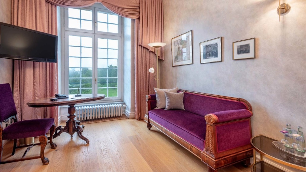 Suite mit Sofa Elegante Suite mit lila Sofa, Holztisch, Stuhl und großem Fenster mit Blick ins Grüne