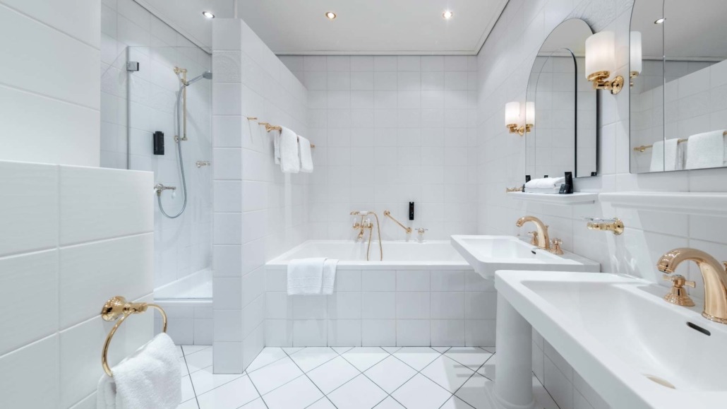 Suite - Badezimmer Helle Suite-Badezimmer mit Badewanne, zwei Waschbecken mit goldfarbenen Armaturen und großer Spiegelwand