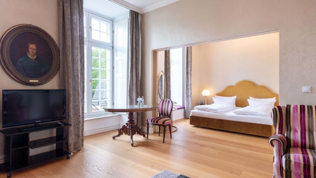 Suite Wohnraum Suite mit großem Fenster, Doppelbett mit gelbem Kopfteil, runder Holztisch und gestreiften Stühlen