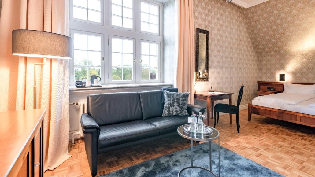 Suite Wohnraum mit Bett Suite mit schwarzem Ledersofa, großem Fenster, Holzparkett, Schreibtisch mit Stuhl und Doppelbett mit Nachttisch