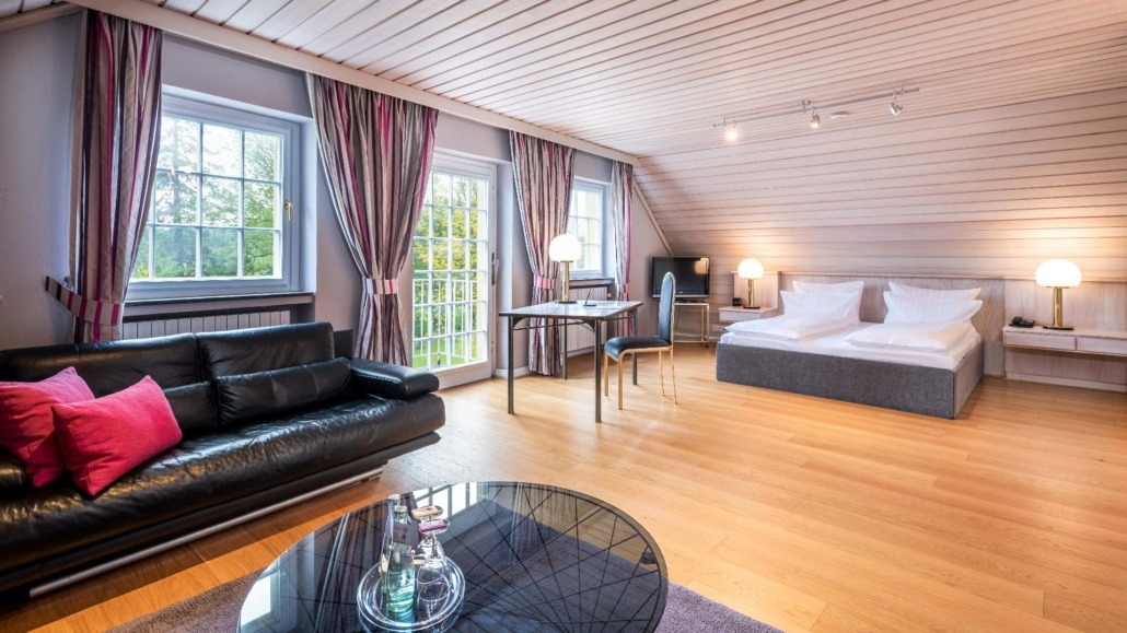 Große Suite Suite mit großem Bett, schwarzem Ledersofa mit roten Kissen, Holzboden und Schreibtisch vor zwei Fenstern mit Vorhängen