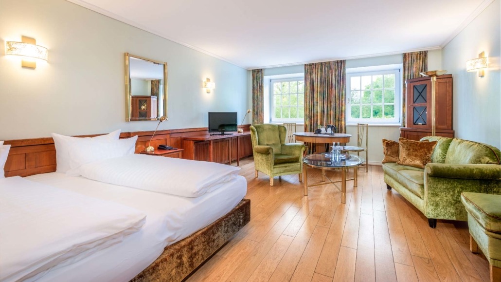 Suite mit kleinem Wohnraum Suite mit großem Bett, grünem Sessel und Sofa, Holzboden, zwei Fenstern mit Vorhängen und einem kleinen Tisch mit Stühlen