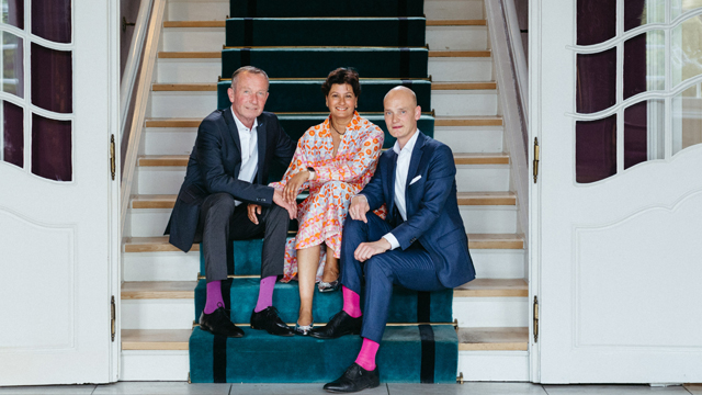 Drei Personen der Familie Winnecken stehen auf einer weißen Brücke im Freien, zwei Männer in Anzügen und eine Frau in einem gemusterten Kleid.